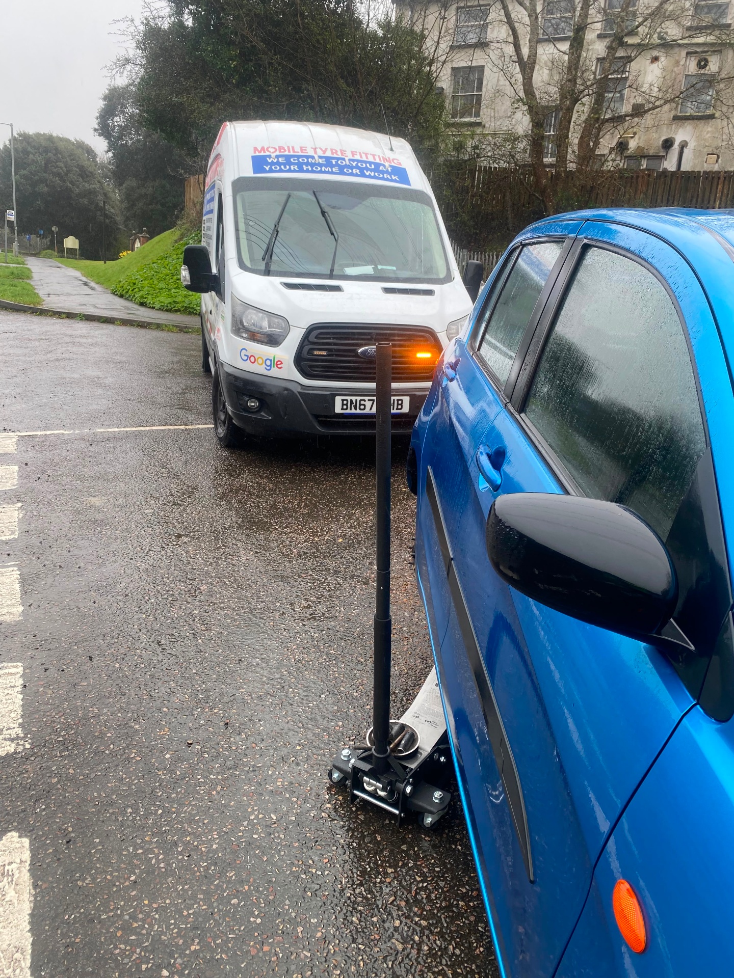Mobile tyre fitting in wet weather — jack under a blue hatchback with Mobile Tyre Fitter 247 van behind